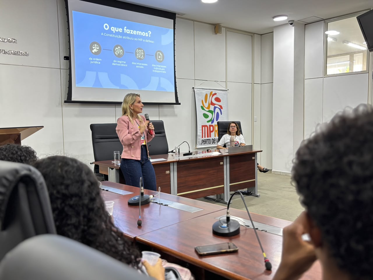 Em auditório, uma mulher loira de casaco rosa palestra em pé para uma plateia vista de costas em primeiro plano, com projeção ao fundo com o título "O que fazemos?" e quatro ícones explicativos. À direita, atrás da bancada, outra mulher branca de óculos e camisa branca está sentada, próxima a um banner com o logo do MPRN e a frase "PERTO DE VOCÊ".