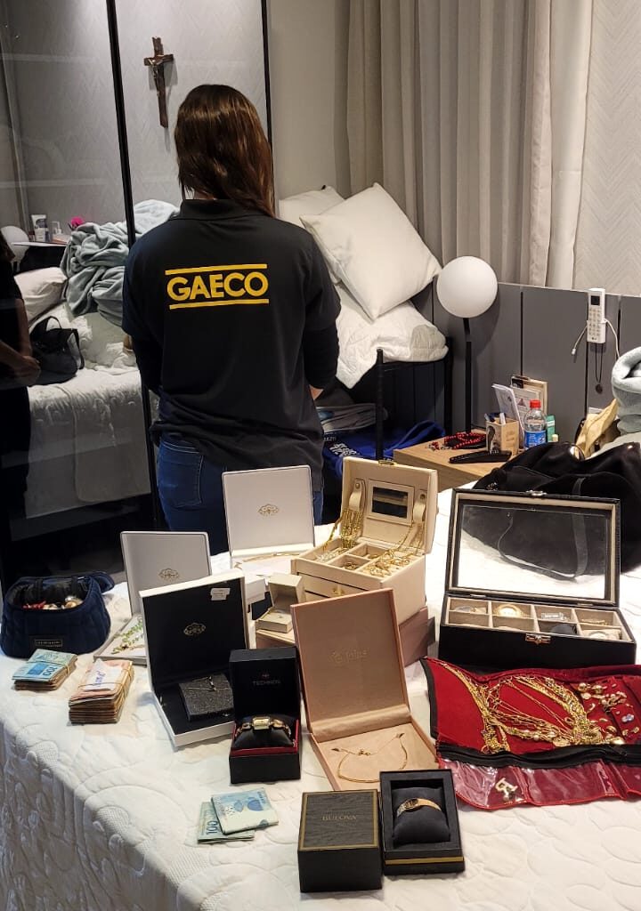mulher de costas ao lado de uma cama com varios caixas de joias e relogios de luxo