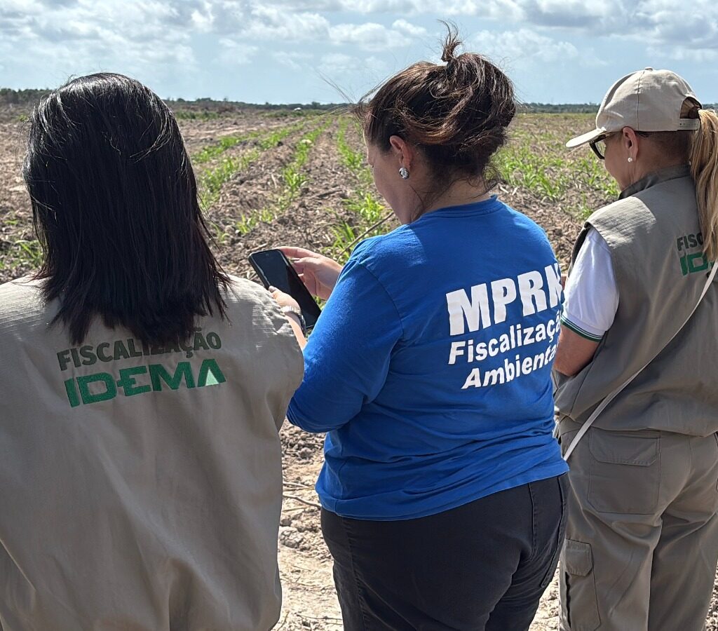 3 mulheres de costas olhando para uma area desmatada, duas delas usam colete do idema e a do meio usa uma blusa escrito MPRN
