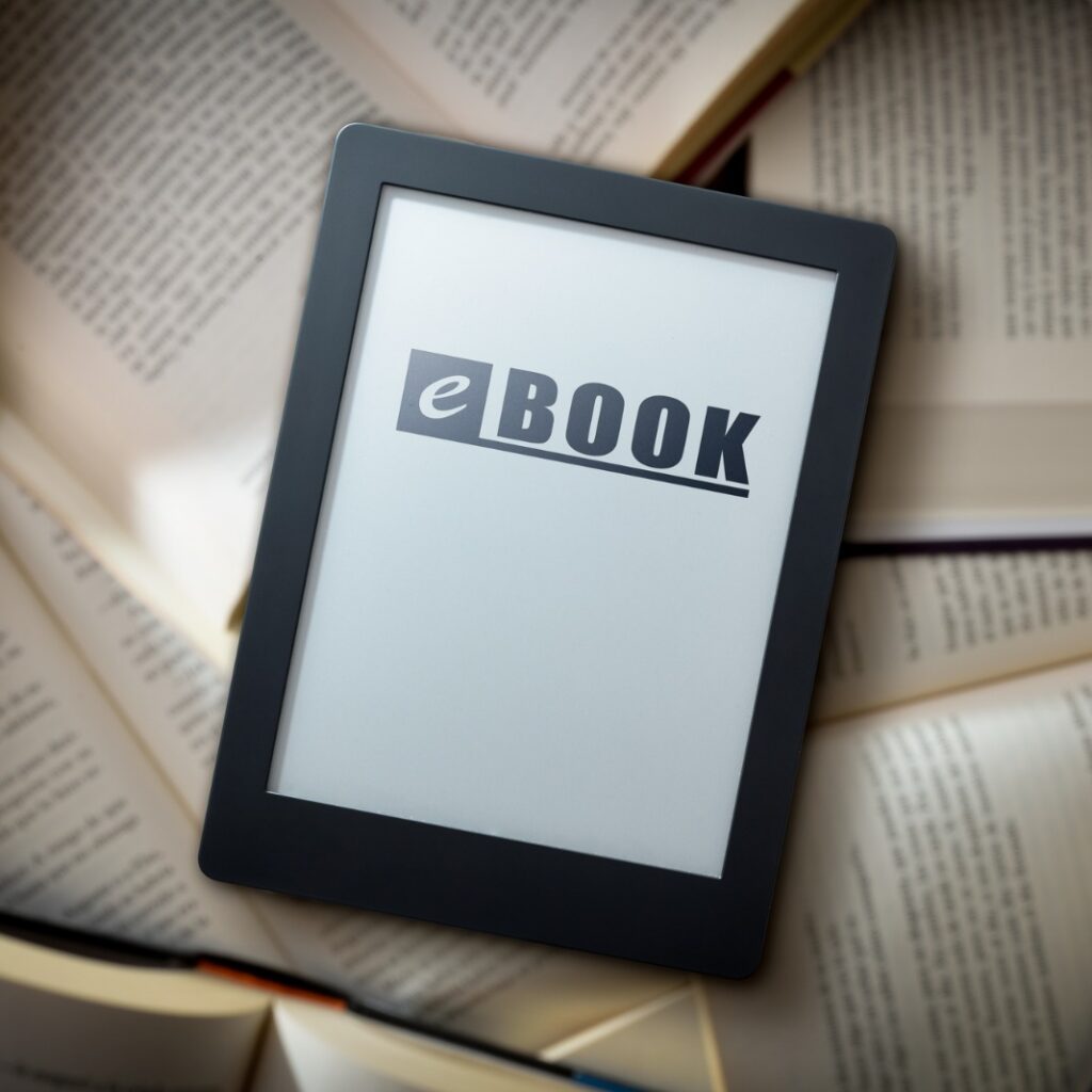 Leitor digital com a palavra eBOOK escrita na tela, posicionado sobre uma pilha de livros físicos abertos.