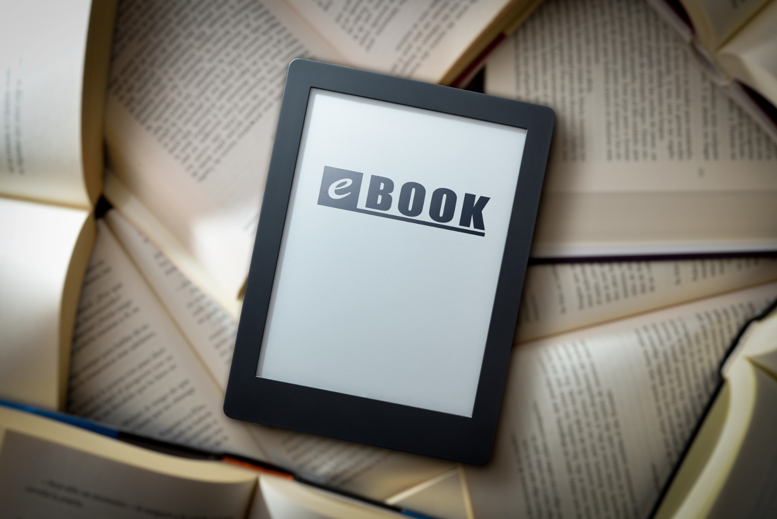 Leitor digital com a palavra eBOOK escrita na tela, posicionado sobre uma pilha de livros físicos abertos.