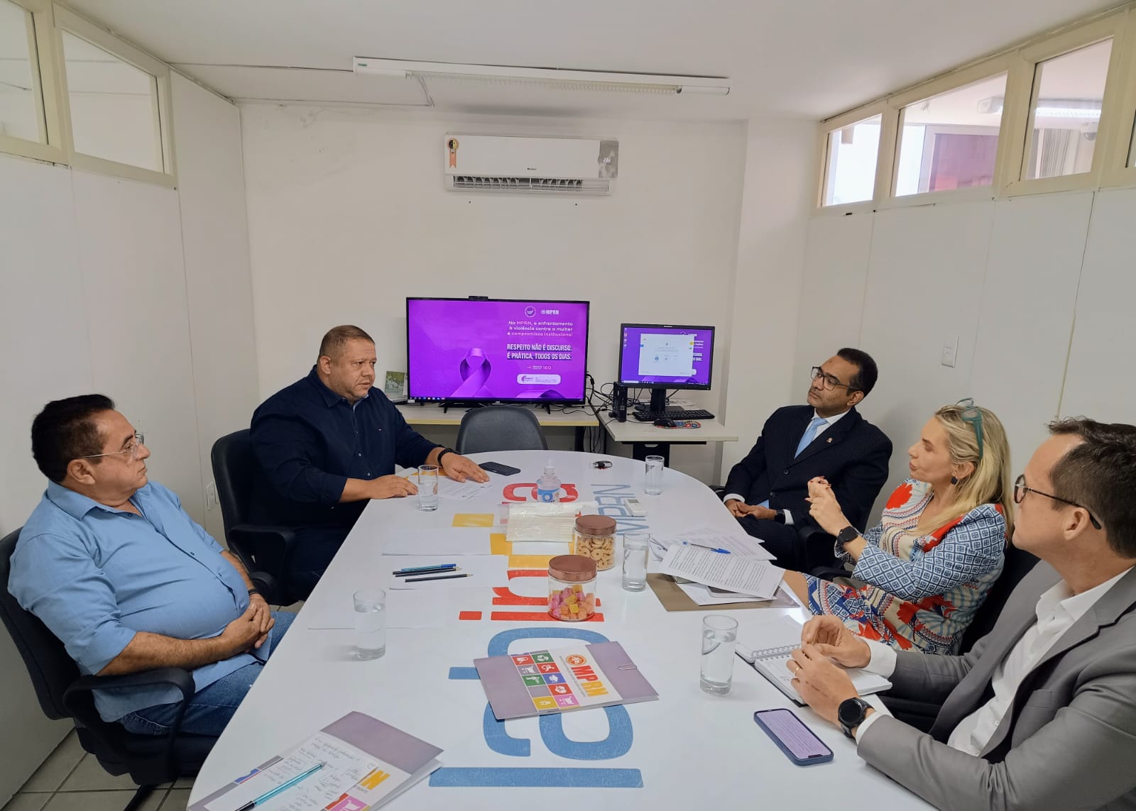 Grupo de pessoas reunido ao redor de uma mesa de reunião em uma sala de escritório, enquanto conversam diante de uma tela ao fundo com uma mensagem roxa de conscientização.