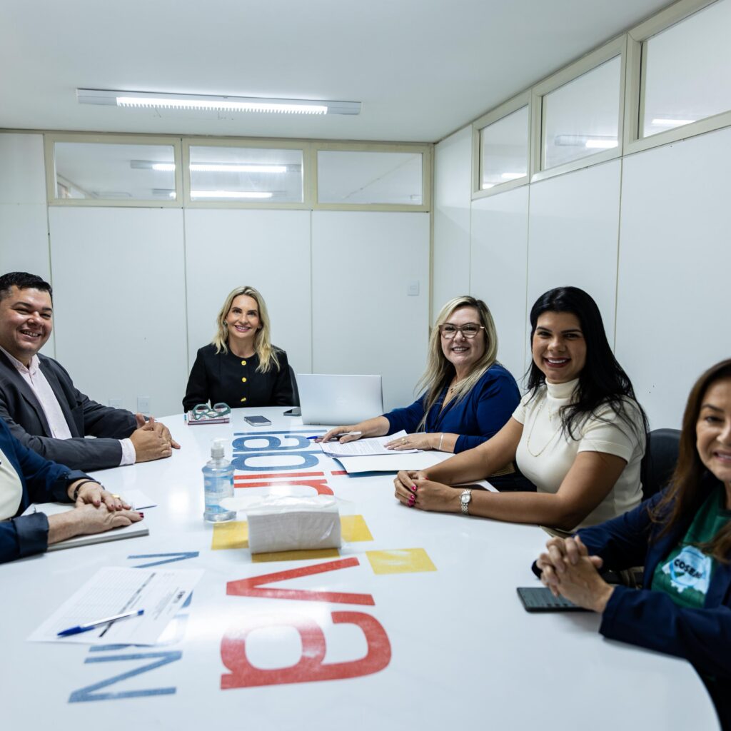Seis pessoas estão reunidas e sorrindo ao redor de uma grande mesa branca de escritório em uma sala de reuniões iluminada.