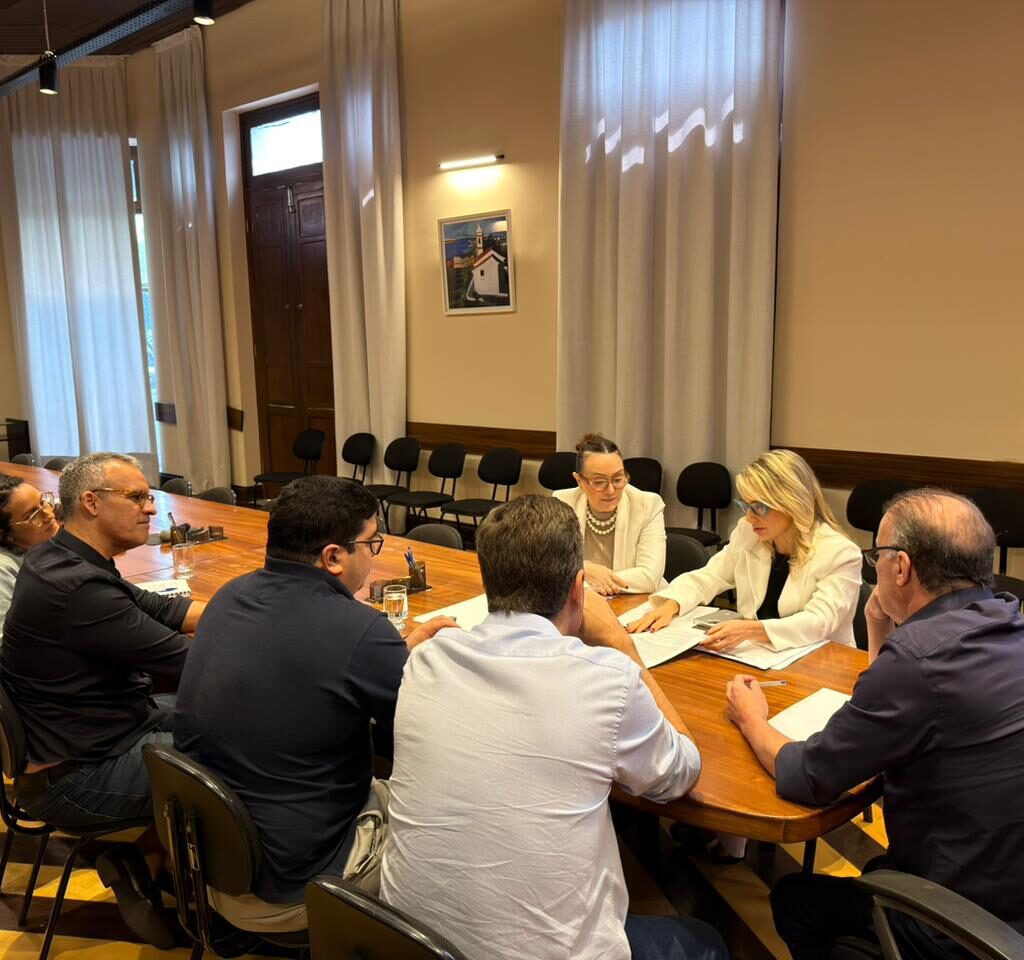 seis pessoas sentadas em mesa de reuniao com o prefeito a ponta e promotoras ao lado