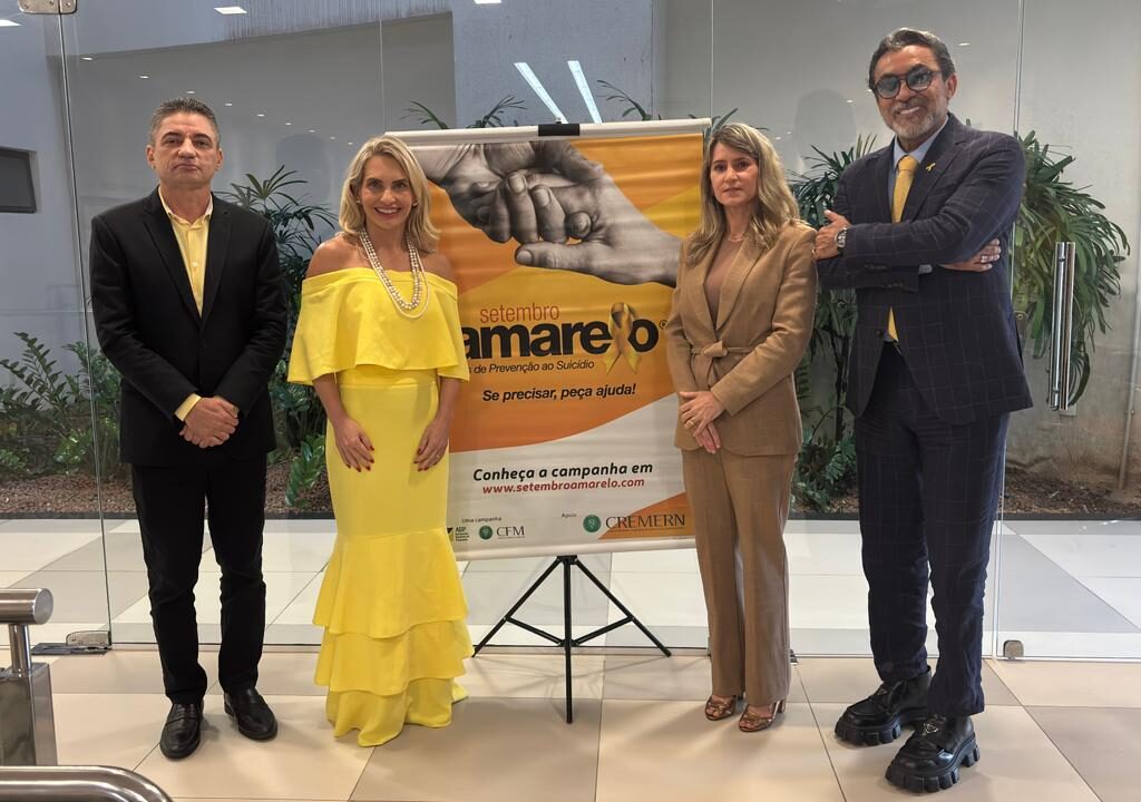 Quatro pessoas vestidas formalmente estão lado a lado ao redor de um banner da campanha Setembro Amarelo.