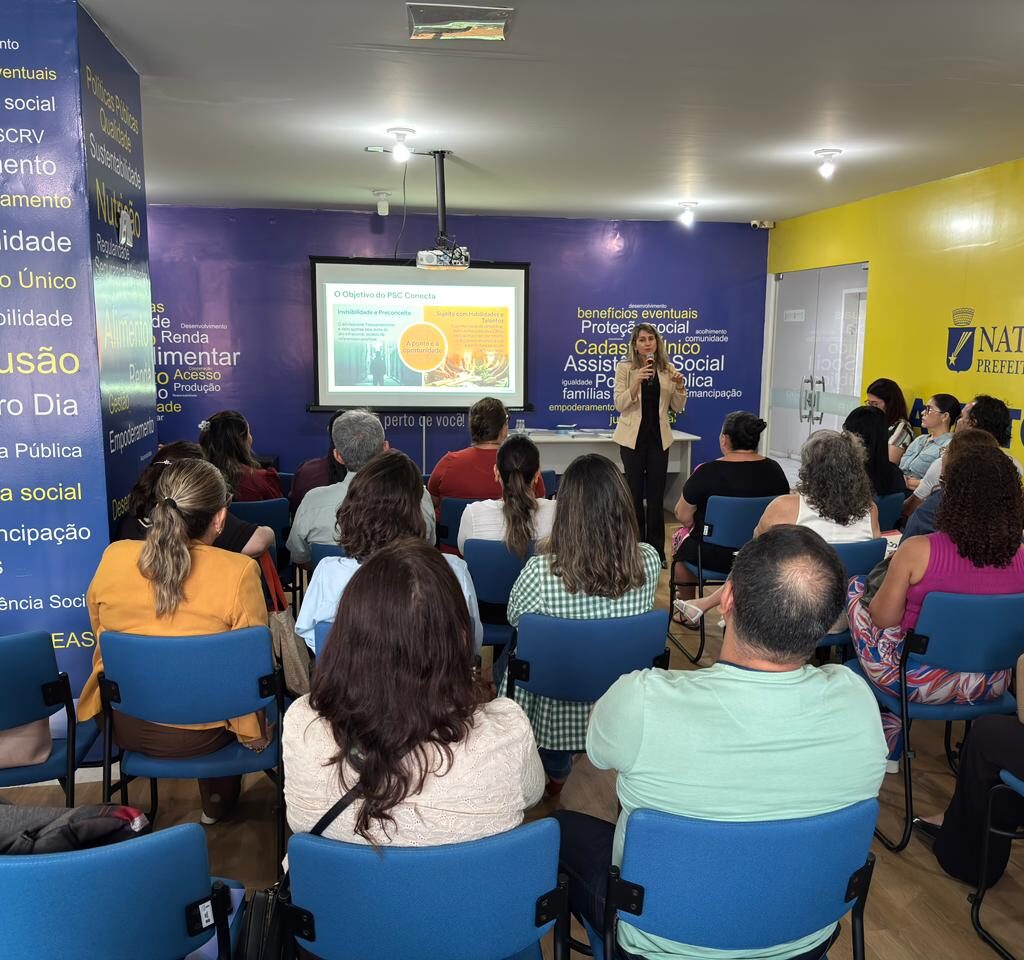 Uma mulher em pé fala ao microfone diante de uma plateia sentada em um auditório colorido, assistindo a uma apresentação de slides.