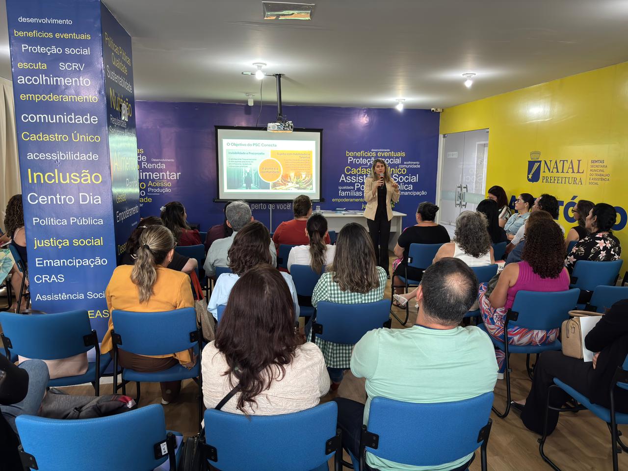 Uma mulher em pé fala ao microfone diante de uma plateia sentada em um auditório colorido, assistindo a uma apresentação de slides.