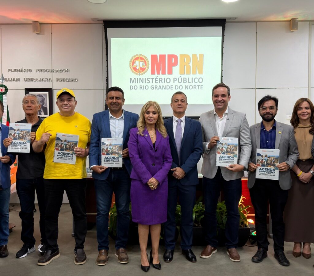 Um grupo de dez pessoas está alinhado lado a lado em um auditório, a maioria segurando livretos, diante de uma tela com o logotipo do MPRN