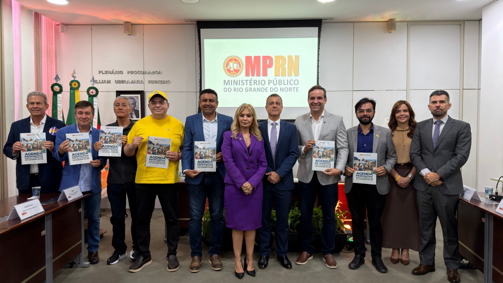 Um grupo de dez pessoas está alinhado lado a lado em um auditório, a maioria segurando livretos, diante de uma tela com o logotipo do MPRN
