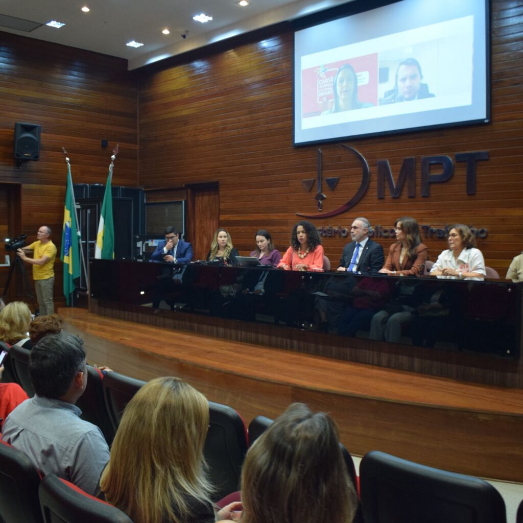 Pessoas assistem a uma apresentação em um auditório com paredes de madeira, onde palestrantes estão dispostos em uma mesa no palco em frente ao logotipo do MPT.