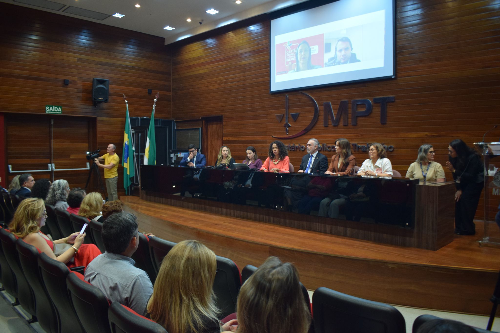 Pessoas assistem a uma apresentação em um auditório com paredes de madeira, onde palestrantes estão dispostos em uma mesa no palco em frente ao logotipo do MPT.