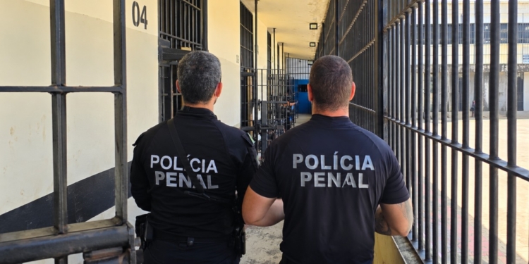 Dois policiais penais de costas em um corredor de um presídio em frente a celas com grades de ferro.