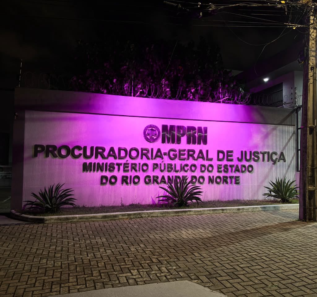 Fachada do prédio da Procuradoria-Geral de Justiça do Ministério Público do Rio Grande do Norte iluminada por luzes cor-de-rosa durante a noite.