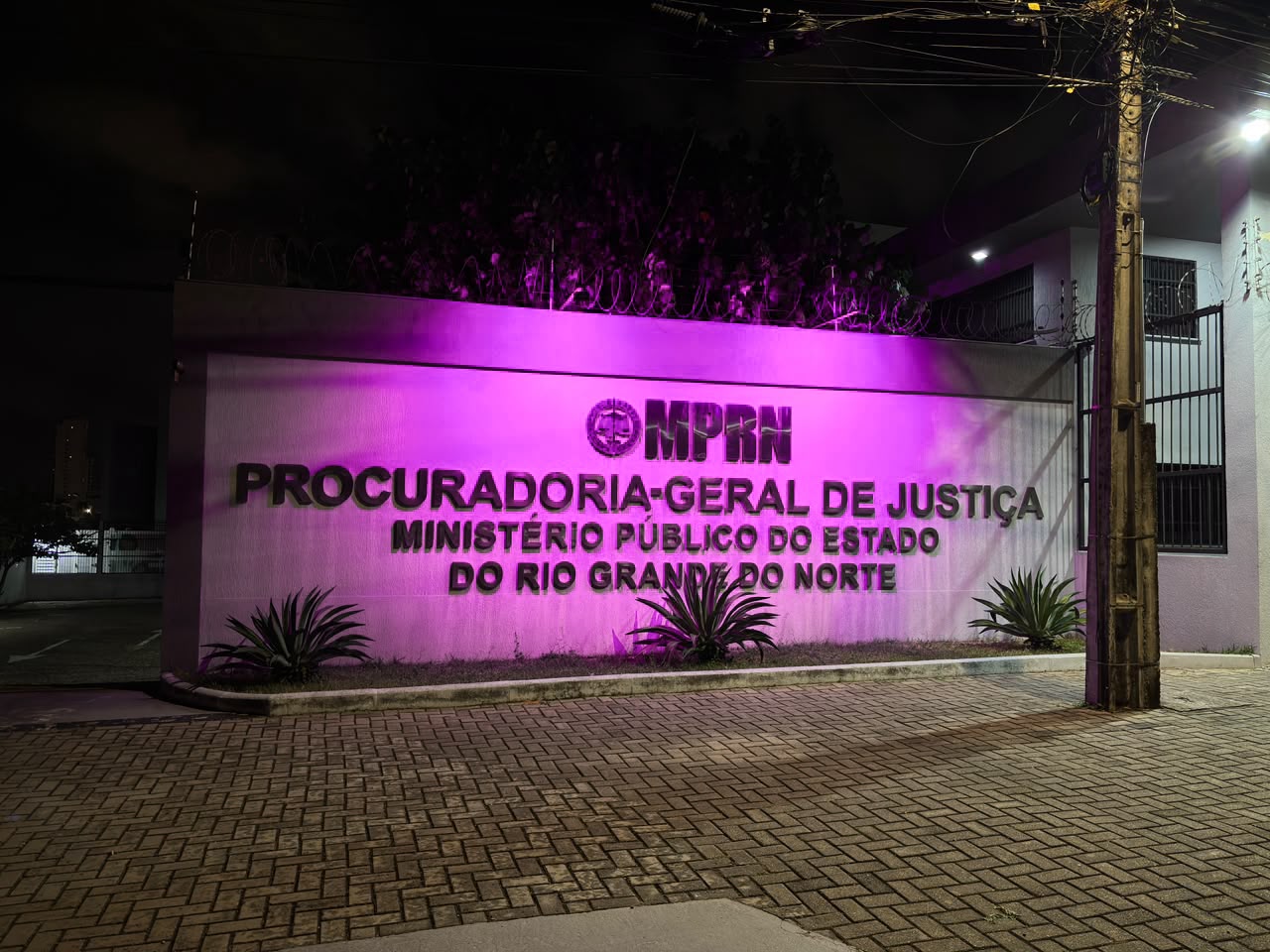 Fachada do prédio da Procuradoria-Geral de Justiça do Ministério Público do Rio Grande do Norte iluminada por luzes cor-de-rosa durante a noite.