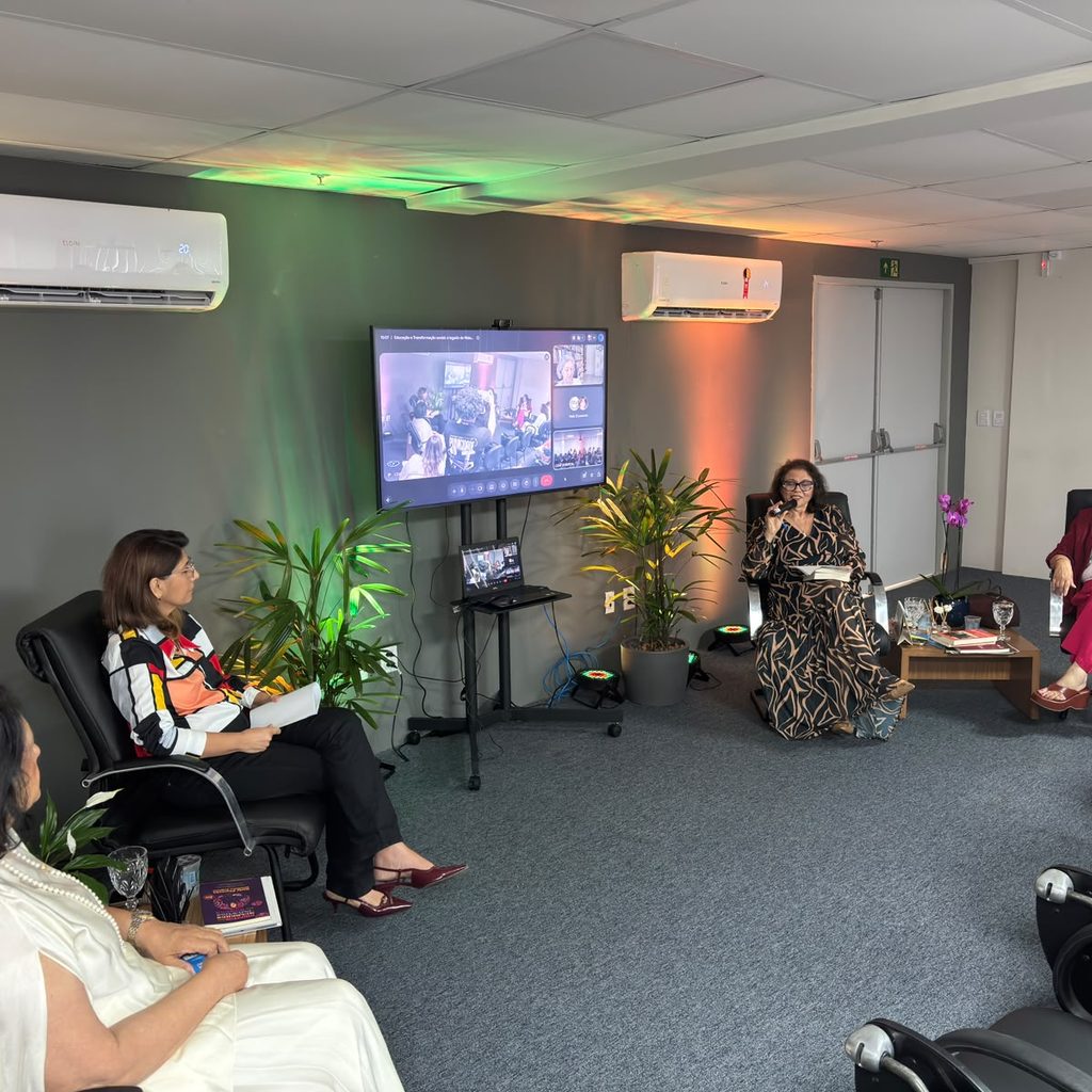 Quatro mulheres participam de uma roda de conversa em uma sala com poltronas e plantas, ao lado de uma tela de televisão que exibe outros participantes por videochamada