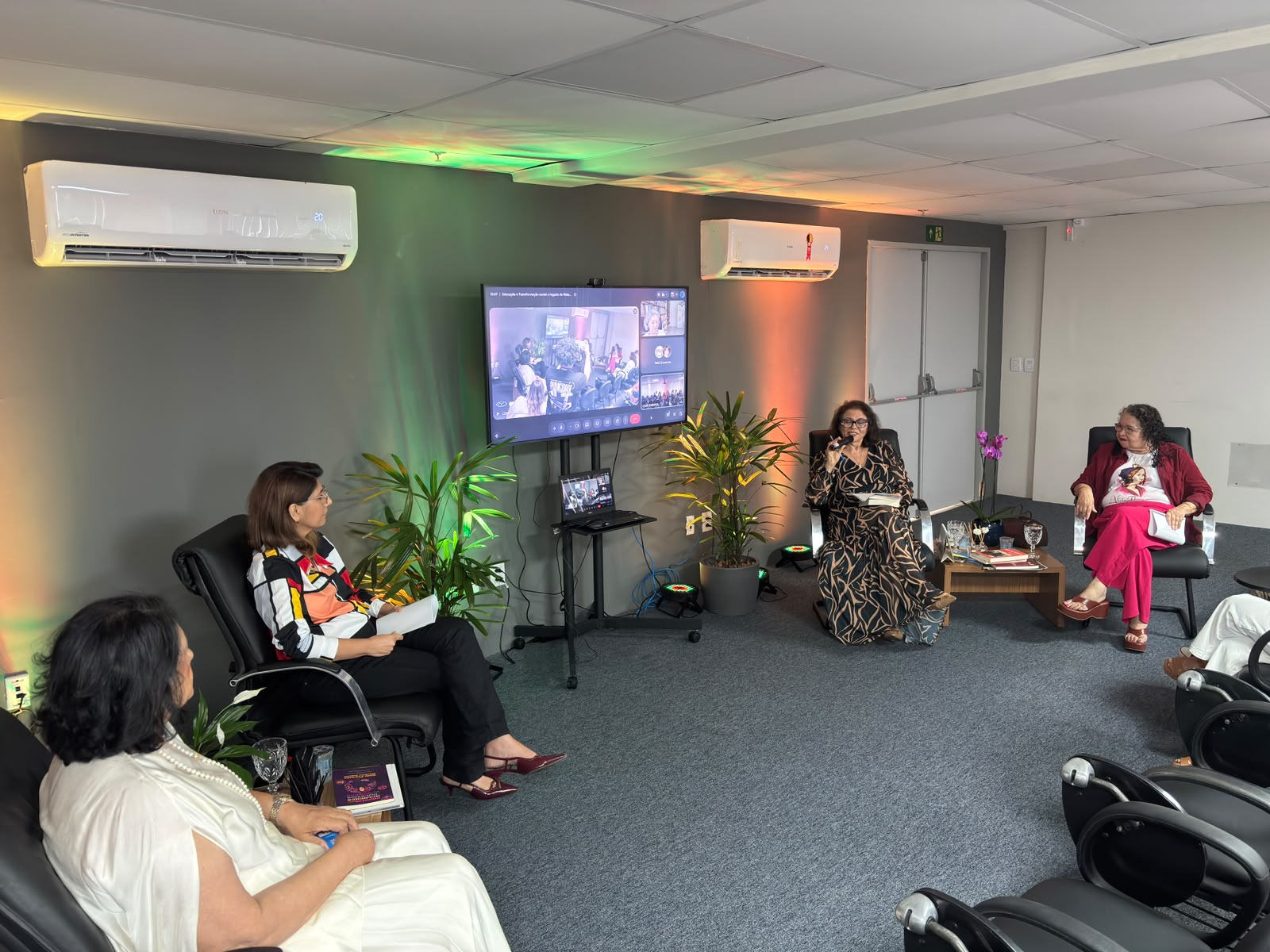Quatro mulheres participam de uma roda de conversa em uma sala com poltronas e plantas, ao lado de uma tela de televisão que exibe outros participantes por videochamada