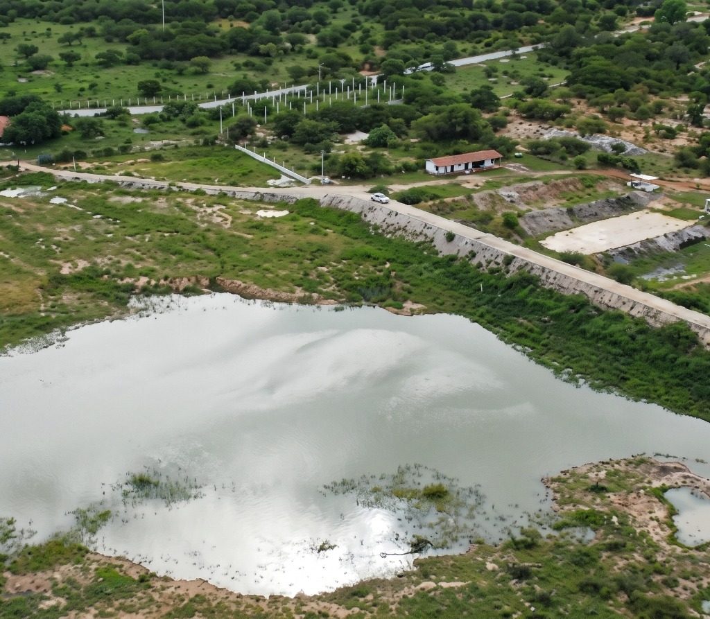 Vista aérea de um lago refletindo o céu cercado por vegetação verde, com uma longa estrada passando ao lado e algumas construções rurais ao fundo.