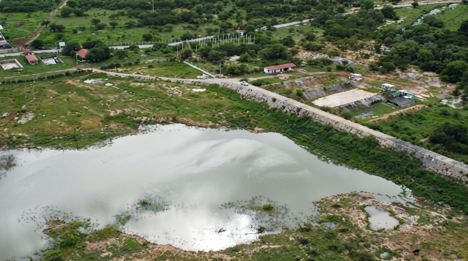 Vista aérea de um lago refletindo o céu cercado por vegetação verde, com uma longa estrada passando ao lado e algumas construções rurais ao fundo.