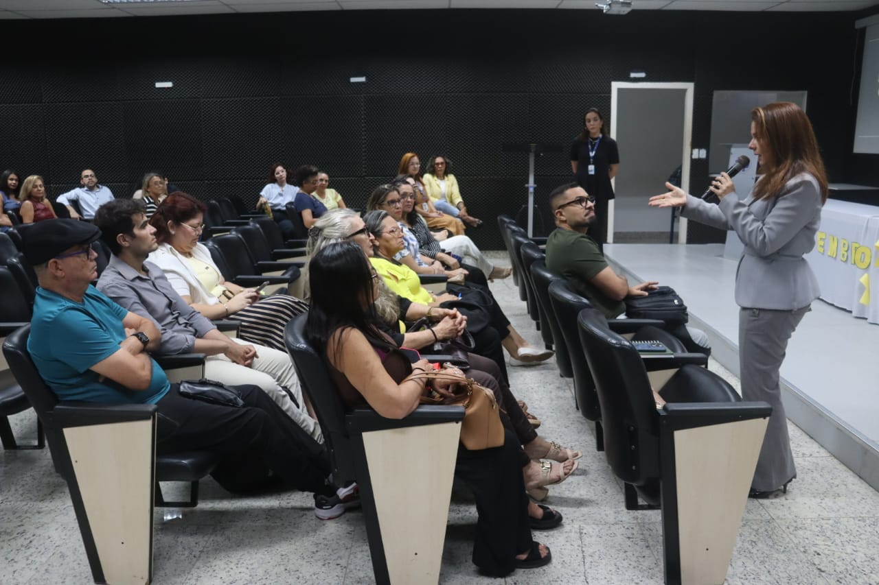 Uma mulher em terno cinza segura um microfone e fala para uma plateia sentada em um auditório.
