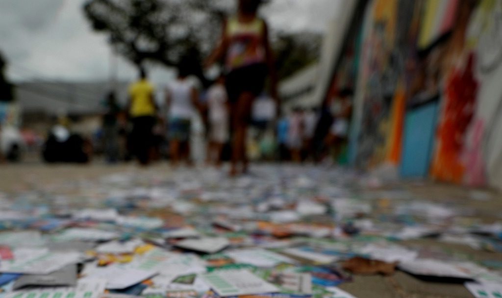 rua urbana com o chão completamente coberto por santinhos e panfletos de campanha eleitoral.